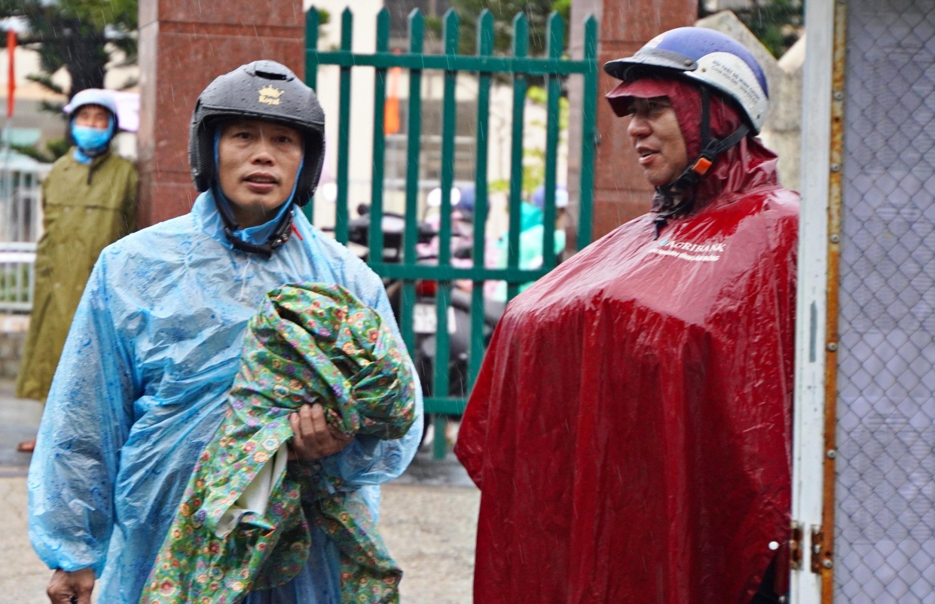 Hai phụ huynh ôm áo mưa đợi con trước cổng trường thi..jpg